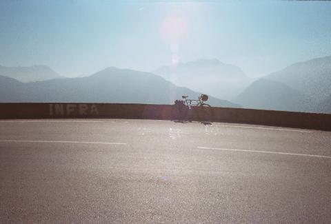 Im vordergrund Straße, von oben strahlt die Sonne, in der Mitte eine Begrenzung vor der ein Fahrrad mit Gepäck steht, dahinter weit entfernte Berge im Dunst.