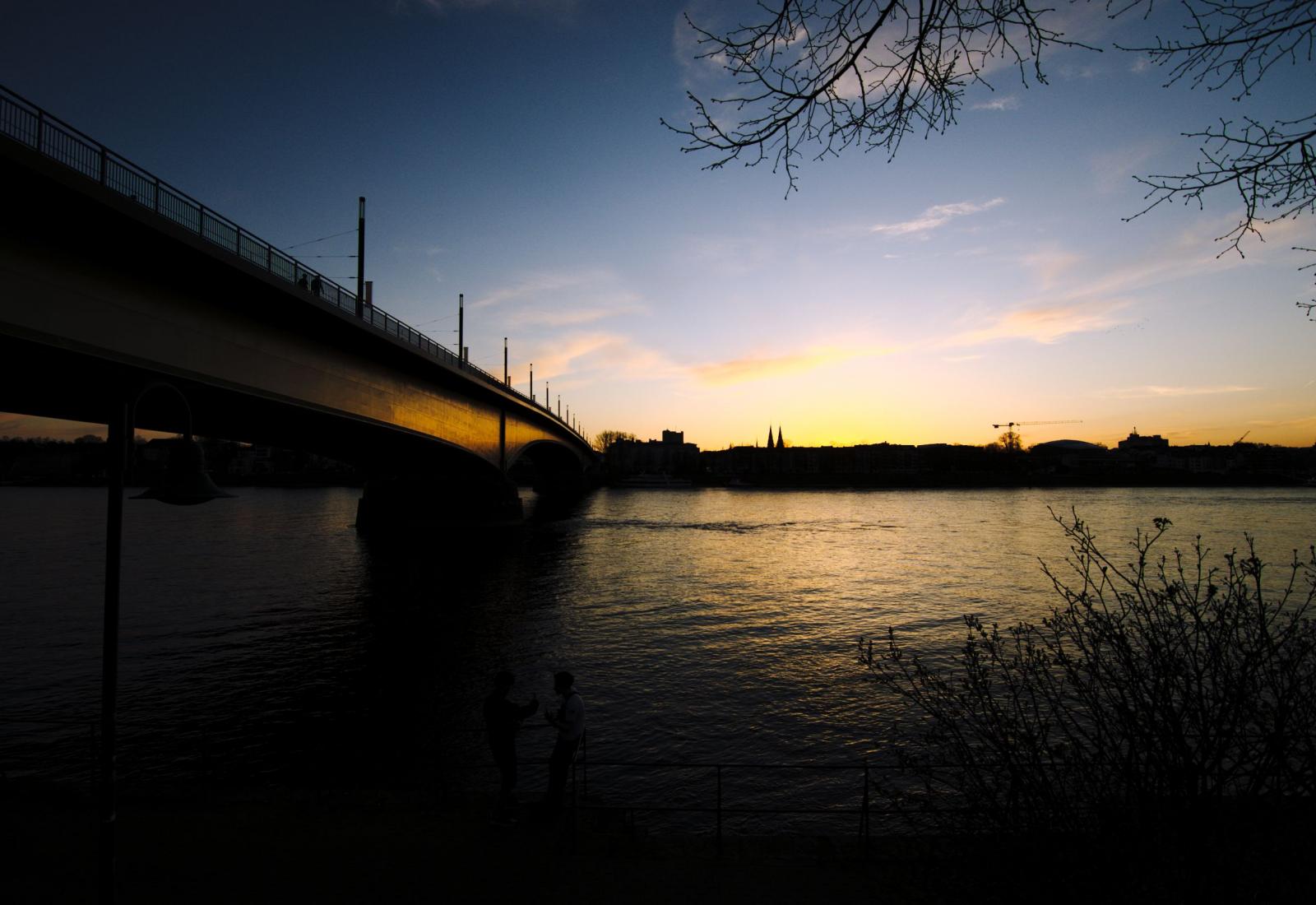 Links die Kennedy Brücke mit Reflektionen der Sonne, rechts oben und unten karge Äste in der Dämmerung