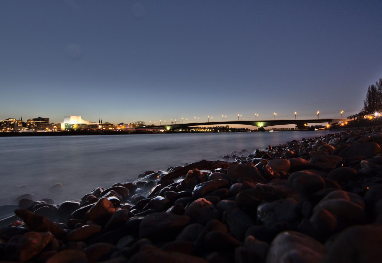 Rechts und unten Steine sehr nach, links mittig der Rhein mit glatter Wasseroberfläche, im Hintergrund rechts die beleuchtete Kennedybrücke und links die beleuchtete Oper im Halbdunkel