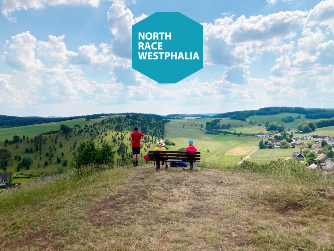 Ausblick auf spärlich bewaldete Hügel, davor 3 Menschen auf und neben einer Bank. Darüber eingeblendet das NorthRaceWestphalia Logo