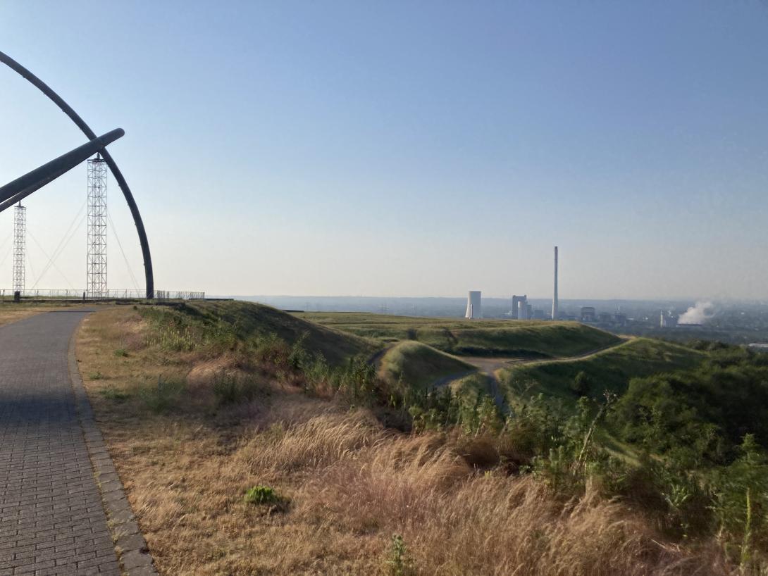 Links ein rundes großes Stahlkonstrukt, rechts der Blick von der Halde ins Ruhrgebiet