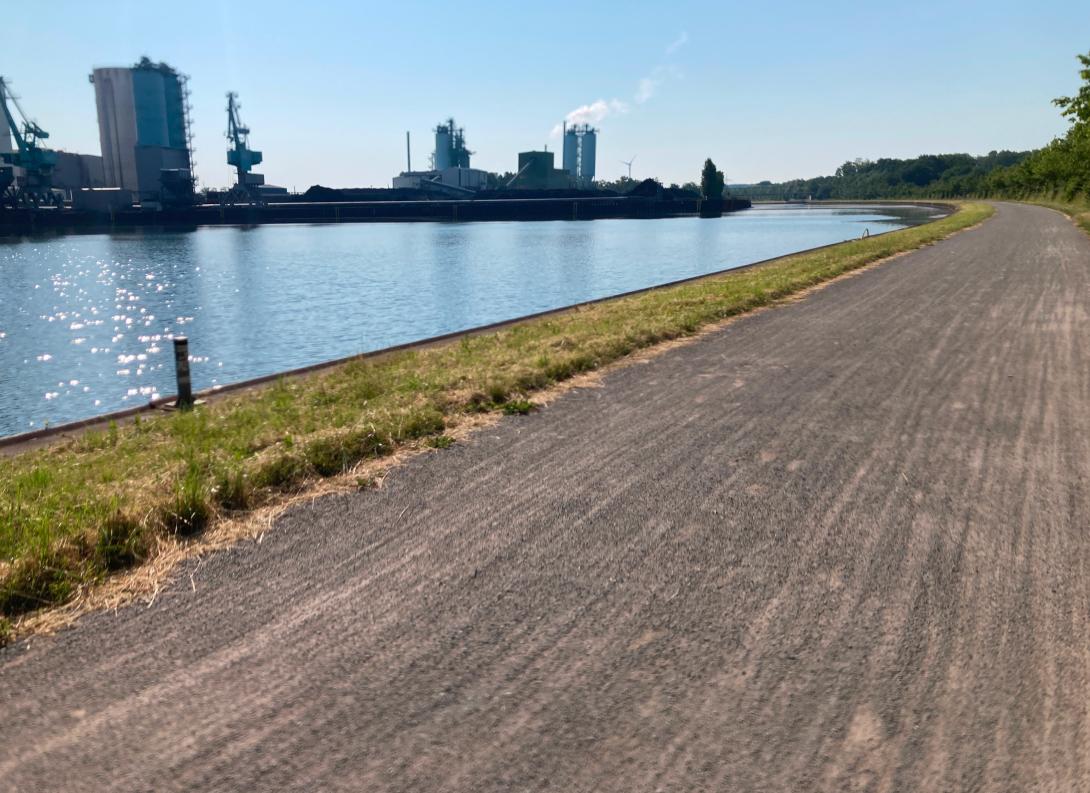 Schotterweg, links daneben ein Fluss und dahinter Industriegebäude