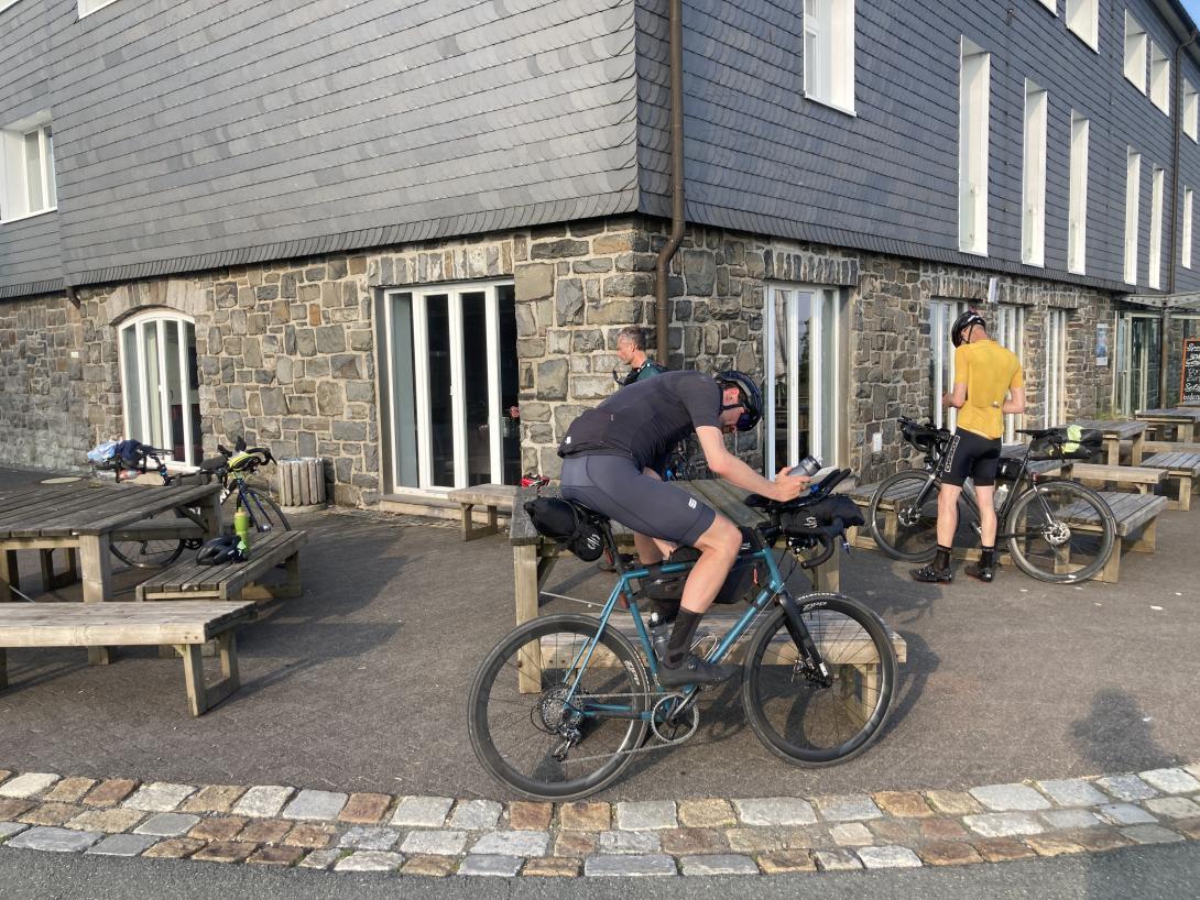 Drei Radfahrer die sich versorgen, dahinter Bänke und ein Gebäude