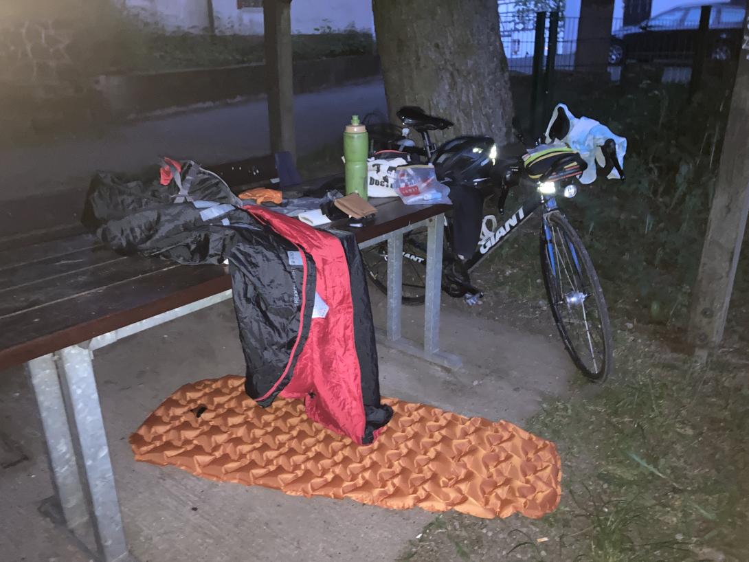 Isomatte unter einem Tisch, da drauf ein Schlafsack und andere Dinge, daneben ein Rennrad